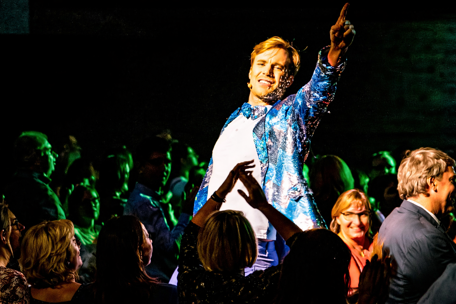 Dynamische Grease-Szene: Ein Paar in Lederjacke und Rock-Outfit im leidenschaftlichen Tanz, umgeben von fröhlichen Tänzern in typischen 50er-Jahre-Looks – vor knallbunter Kulisse mit rotem Oldtimer!