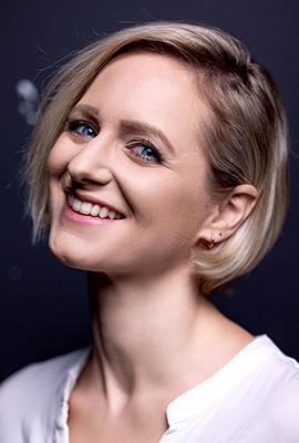 Das Portrait zeigt die Darstellerin Lina Ritters mit kurzem, blondem Haar und einem strahlenden Lächeln. Sie trägt ein helles Oberteil, der dunkle Hintergrund hebt ihr Gesicht hervor.