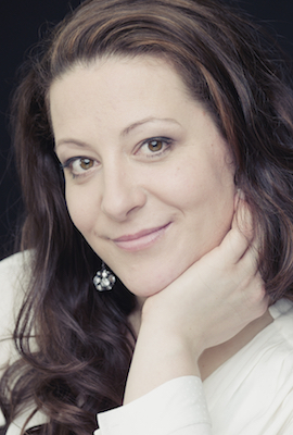 Das Bild zeigt Nadine Hammer, Darstellerin bei Best of Musicals, mit langen, gewellten braunen Haaren und einem freundlichen Lächeln. Sie trägt ein rotes Oberteil, einen schwarzen Blazer und große Ohrringe.