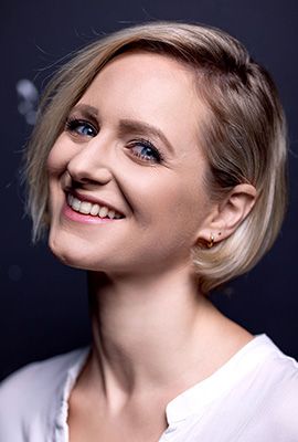 Das Portrait zeigt die Darstellerin Lina Ritters mit kurzem, blondem Haar und einem strahlenden Lächeln. Sie trägt ein helles Oberteil, der dunkle Hintergrund hebt ihr Gesicht hervor.